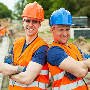 Two smiling construction workers in hard hats (orange and blue) and safety vests, arms crossed.